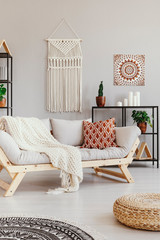 Blanket and cushion on couch in boho living room interior with pouf and poster. Real photo