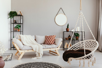 Hanging chair near grey wooden sofa in ethno living room interior with round mirror. Real photo