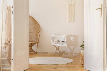 Open door to white scandi kid room with crib and wicker peacock and carpet on the parquet, real...