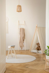 Vertical view of white scandinavian playroom with tent, teddy bear and wooden ladder with beige blanket, real photo