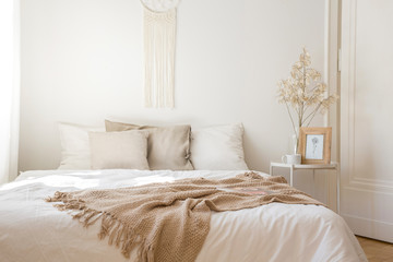 King size bed with white bedding, beige pillows and blanket next to bedside table with flower,...