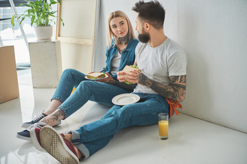 young tattooed couple eating sandwiches and looking at each other while sitting on floor i new house
