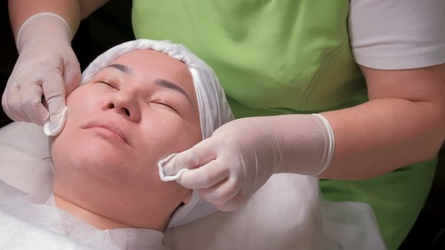 Beautician in a green dress wipes the face of an oriental girl with cotton pads. An asian muslim woman in a beauty salon is lying during a rejuvenating skin procedure. Women's hands in white gloves.