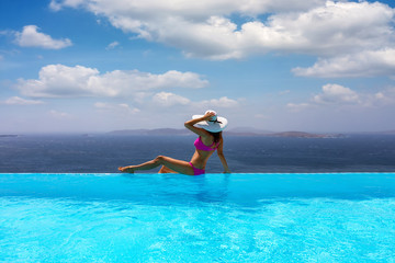 Attraktive Frau im Bikini und mit weißem Hut genießt die Aussicht von einem Pool auf das Meer 