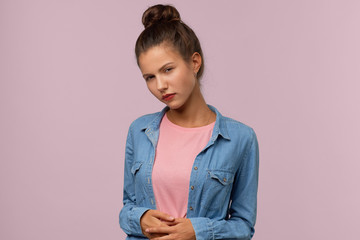Obraz premium A beautiful brunette girl with a bun of hair on her head frowns and holds her stomach. Studio portrait on isolated background. The concept of irritability, frustration and discomfort in the abdomen