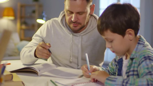 Rack focused shot of Caucasian man reading textbook and talking to son while helping kid with study at home