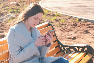 Portrait of beautiful brunette reading and typing a message in her phone sitting on the bench in park.