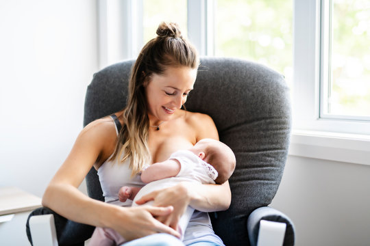 Mother is breastfeeding a baby sitting on a chair close to the window