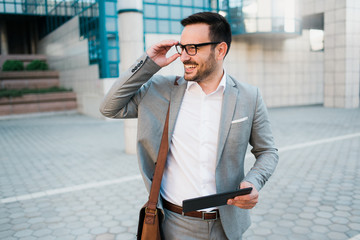Young successful happy businessman using tablet and smiling, wearing elegant suit, business bag and modern glasses. 