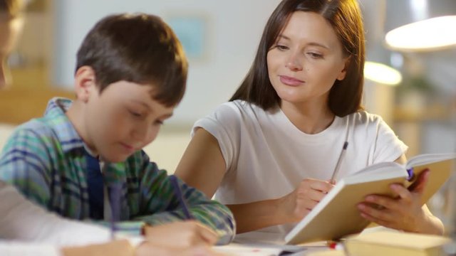 Close-up rack focused shot of two kids doing homework together with mother: brother and sister writing down in notepads while woman reading textbook aloud