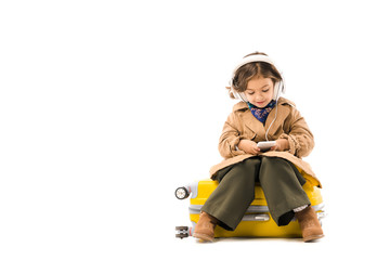 adorable little child in trench coat listening music with headphones and using smartphone while sitting on yellow suitcase isolated on white