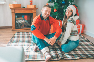 Picture of happy young couple sitting together on blanket. They smile and look at camera. Young men wear sweater. Woman has Christmas hat.