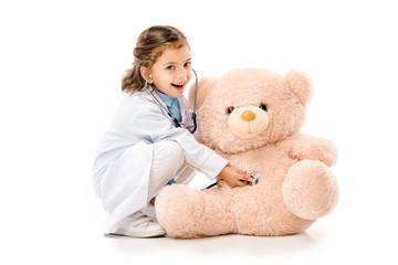 kid dressed in doctors white coat with stethoscope playing with teddy bear isolated on white
