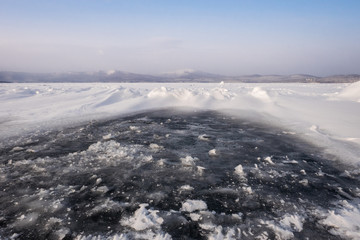 Obraz premium Natural winter lake ice breaking with clear sky background, selective focus. Frozen mountain lake with blue ice and cracks on the surface. Winter landscape with snowy hills under a blue sky.