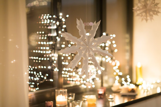 Christmas And Holidays Concept - Paper Snowflake Decoration Hanging Over Garland Lights And Candles At Night Window