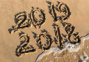 New year 2019 concept. Inscription 2018 and 2019 on a beach sand, sea wave covering digits 2018.