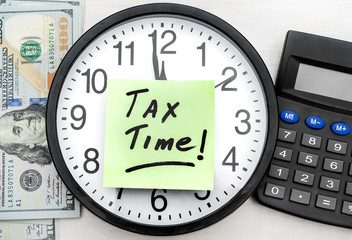 Sticker with phrase "TAX TIME" on clock, calculator and money on wooden background.