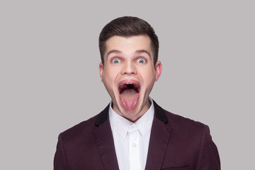 Portrait of funny crazy handsome young man in violet suit and white shirt, standing, looking at camera with open mouth, tongue out and big eyes. indoor studio shot, isolated on grey background.