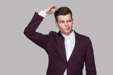 Portrait of thoughtful handsome young man in violet suit and white shirt, standing, looking away and scratching his head with confused face. indoor studio shot, isolated on grey background.