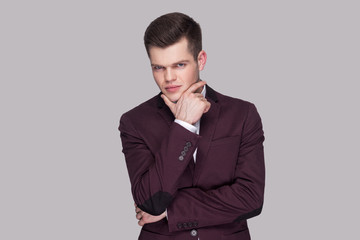 Portrait of handsome serious young man in violet suit and white shirt, standing, touching his chin and looking at camera with thoughtful face. indoor studio shot, isolated on grey background.