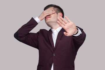 I dont want to see this. Portrait of confused young man in violet suit and white shirt, standing, closed his eyes with hands and showing stop gesture. indoor studio shot, isolated on grey background.