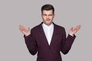 I don't know. Portrait of confused handsome young man in violet suit and white shirt, standing, looking away, raised arms and thinking. indoor studio shot, isolated on grey background.