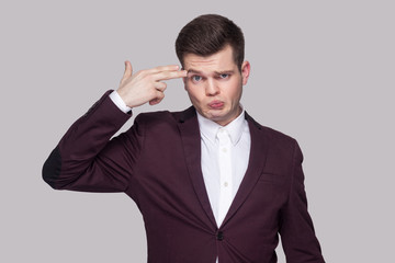Portrait of confused handsome young man in violet suit and white shirt, standing, looking at camera with serious face and gun gesture. indoor studio shot, isolated on grey background.