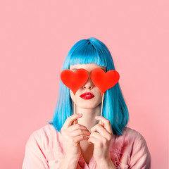 Young stylish woman with blue hairstyle and red lipstick holding valnetine's heart on her eyes.