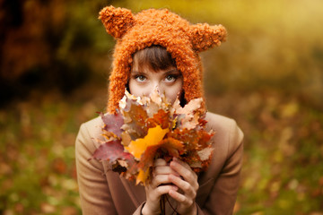 Beautiful happy smiling girl wearing stylish knitted fox hat and camel jacket posing with bunch of mapple leaves in autumn park