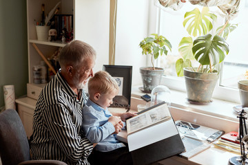 senior man and little boy holding and looking at family photo album in living room