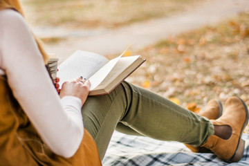 Young girl reading book in park at retro blanket and beautiful autumn day. Woman holds book and coffee cup on legs and wears hot autumn clothes. Close up, selective focus