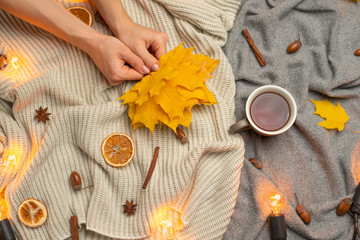 Obraz na płótnie Canvas plaid with hot tea in the hands of the girl and a woolen blanket, autumn yellow leaves and white light bulbs. Conception of warmth, comfort, comfort. View from above