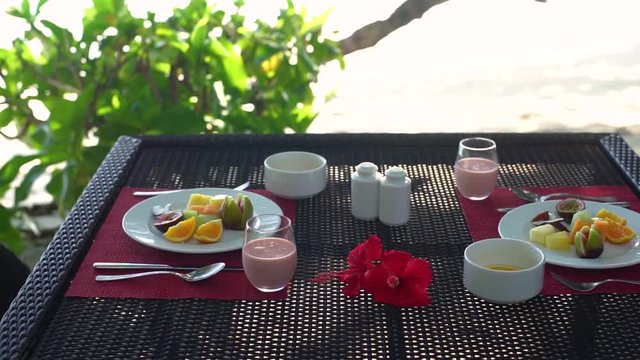 Linear dolly of breakfast table on beach at seaside restaurant, plate with fresh fruit, juice and yoghurt bowls, hibiscus flowers. Slow motion 50p.