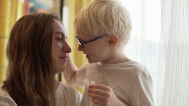 Mom and son are talking. The boy kisses his young mother on the cheek. Beautiful woman smiling. Then he plays each other's noses, kissing like Eskimos. The family is wearing white sweaters.