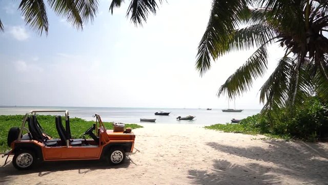 Slow linear dolly shot of tourist beach buggy/Moke parked on tropical island beach, white sand, palm trees and boats on sea. Medium shot, Slow motion 50p.