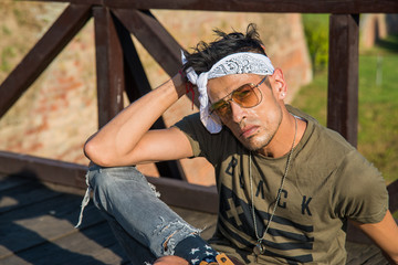 Extravagant (hipster) male model with sunglasses and a white scarf on a wooden bridge.