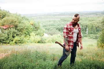 bearded mohawk lumberjack worker standing in forest with axe