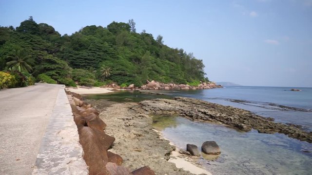 Scenic view of road next to sea shore on tropical island with palm trees as couple drive away with mini Moke/ beach buggy. Seychelles, Praslin. Slow motion 50p.