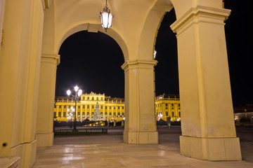 View of illuminated palace in Vienna at night through an archway with pillars.
