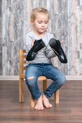 little girl sits on a wooden chair and holds in her hands mother shoes with high heels, studying them