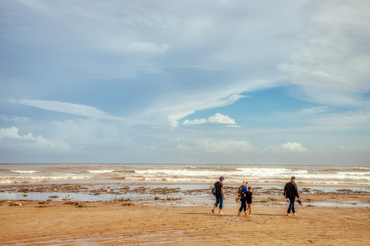 An Unidentifiable Family Walking Along A Littered Beach After A Storm With White Clouds, A Blue Sky, Brown Sand, And Many Waves Crashing On The Shore