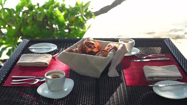 Close-up of breakfast table with cappuccinos, basket of freshly baked croissants as couple (hands) stir coffee, eat pastries, seaside restaurant. Slow motion 50p.