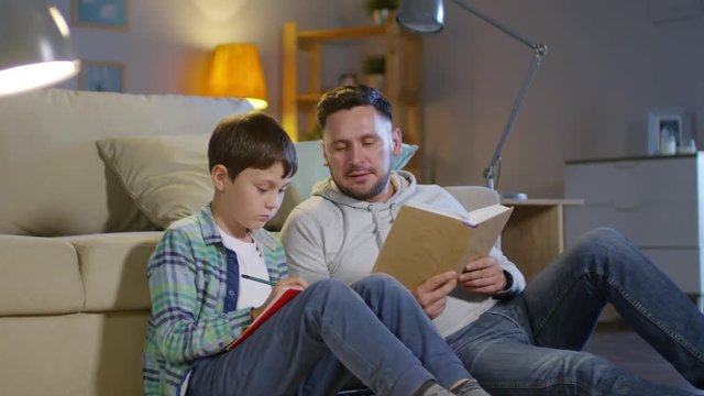 Man sitting on the floor by couch in the living room and reading textbook to little son while doing homework together