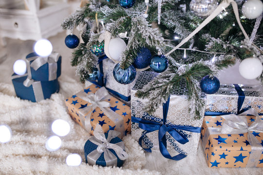 Christmas Gifts Wrapped In Silver And Blue Paper, Background With Xmas Lights Bokeh Of Blurred Under Christmas Tree.