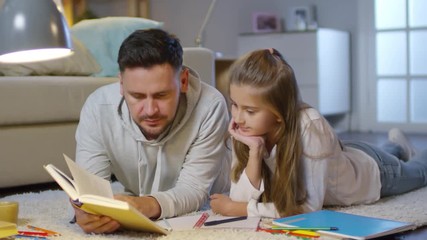 Father lying on comfortable carpet in the living room and reading a book to young daughter while doing homework together at home - Powered by Adobe