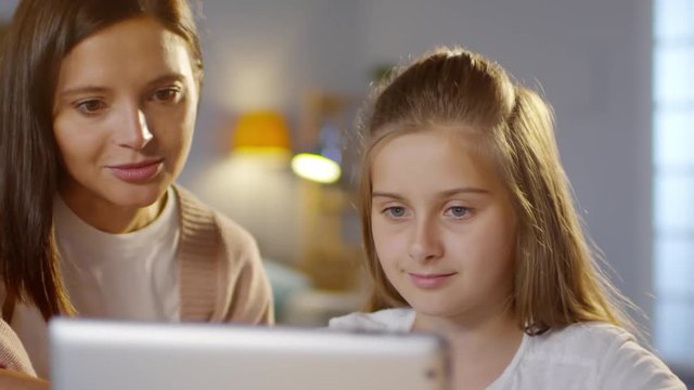 Close-up of beautiful mother and little daughter looking at screen of digital tablet, smiling and talking