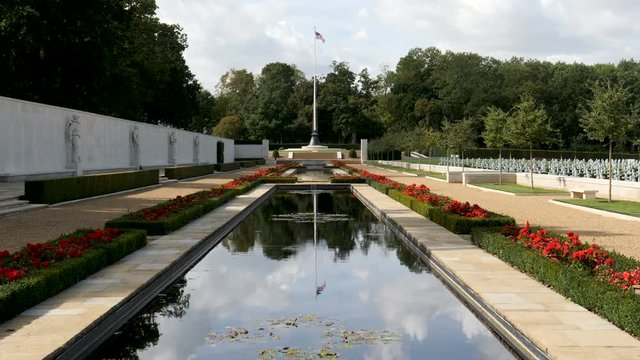 Zoom in-American flag flying over a reflecting pool at the WWII American Cemetery near Cambridge, England with graves of the dead on the right and the Wall of the Missing on the left.