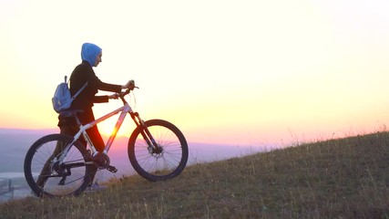 Muslim woman in a hijab with a backpack with a bicycle goes uphill and looks at sunset,slow mo