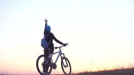 Muslim woman in a hijab with a backpack with a bicycle watching the sunset with her hand up in joy