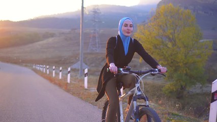 Muslim woman in hijab with backpack riding a bike on the road setting sun